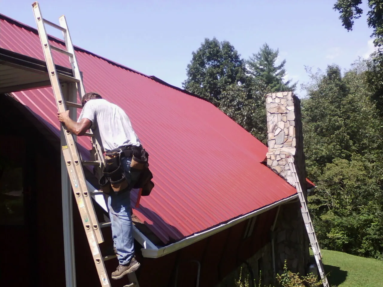 Roofer on a ladder beside a red metal roof and stone chimney — Soffit & Fascia Repair by Roofing Brothers in Gardiner
